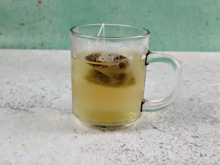 Sachê de chá infusionando em uma xícara de vidro com água gelada.