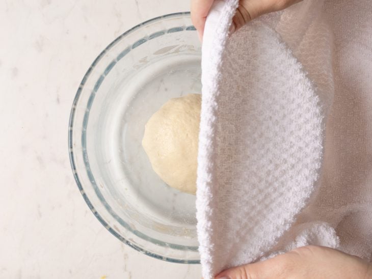 Massa sovada dentro de uma tigela de vidro coberta com um pano úmido.
