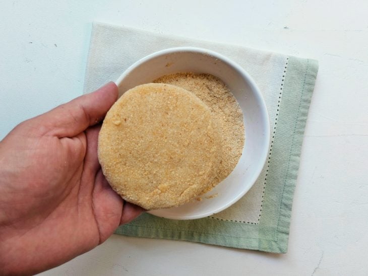 Um hambúrguer cru de frango sendo empanado na farinha de rosca.