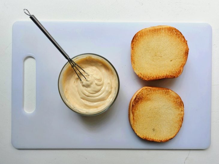 Uma tábua contendo um pão de hambúrguer cortado ao meio e maionese.