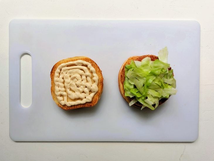 Uma tábua contendo uma metade de pão de hambúrguer com maionese e a outra com hambúrguer de frango e alface.