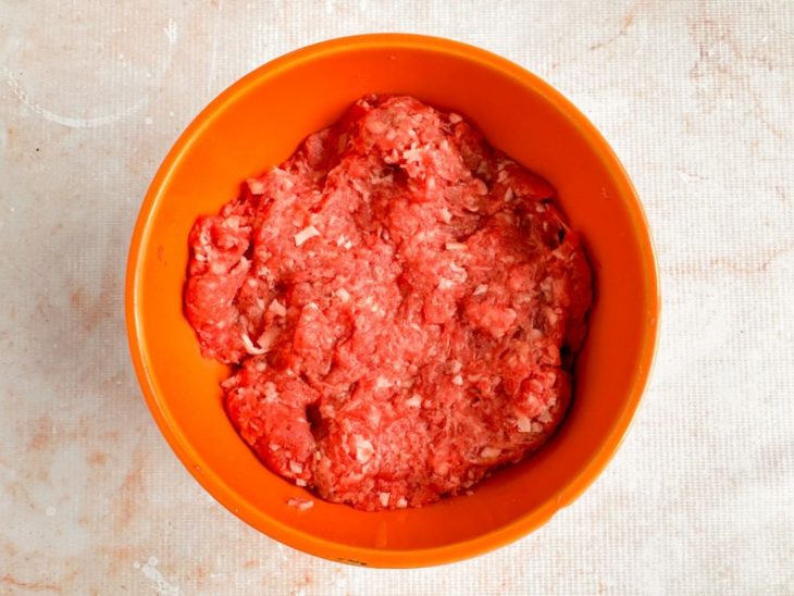 Carnes moídas e bacon sendo misturados com as mãos até formar massa uniforme.