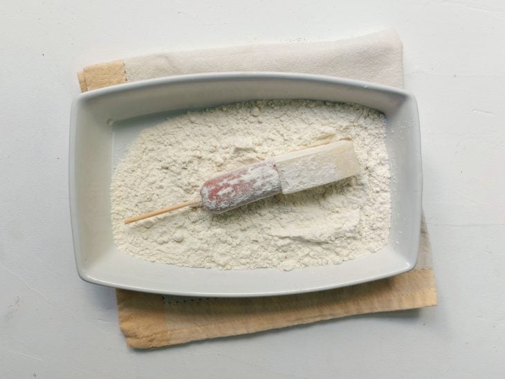 Um recipiente com farinha de trigo e um palito de hot dog sendo empanado,