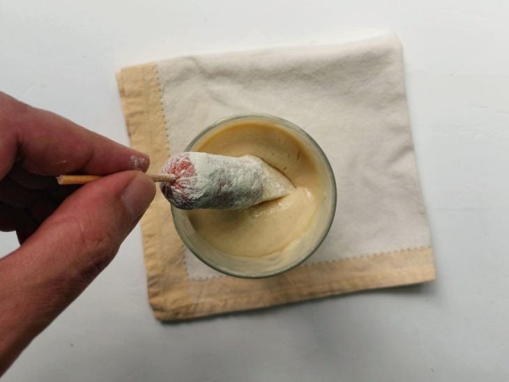 Um palito com salsicha e queijo sendo mergulhado no copo com massa.