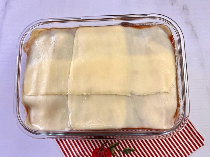 Um refratário forrado com molho sugo e massa de lasanha, coberto com fatias de queijo.