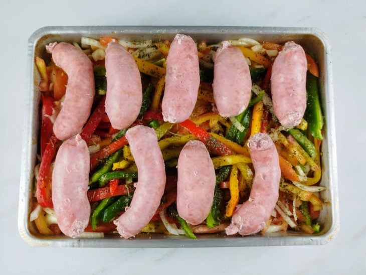 Linguiças, pimentões, tomates, cebolas, azeite, alho, ervas secas, pimenta-do-reino e sal em uma forma de alumínio.