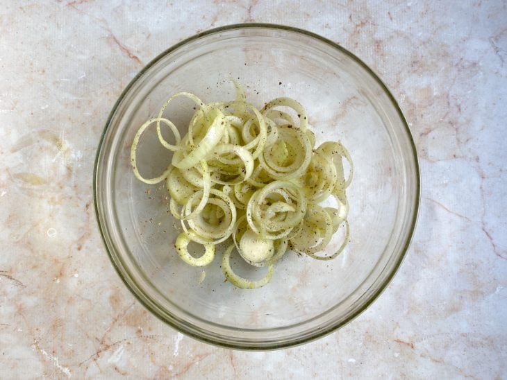 Rodelas de cebola temperadas com azeite, sal e pimenta-do-reino dentro de um recipiente de vidro.