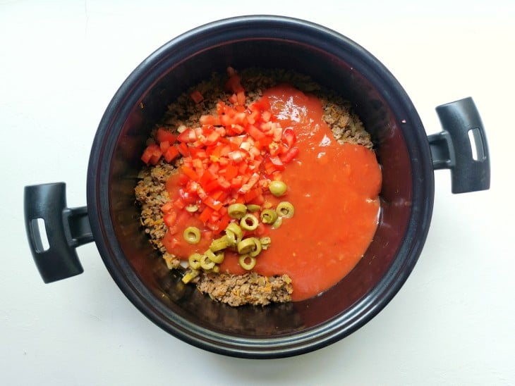 Uma panela contendo cebola, alho, cenoura ralada, carne moída, molho de tomate, tomate e azeitonas.