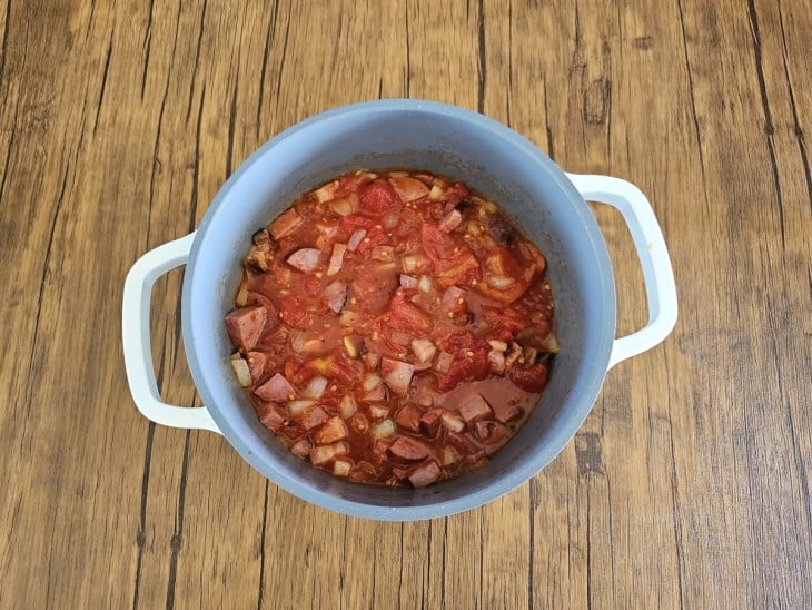 Uma panela de cor cinza com alças na cor branca, cozinhando o ingredientes: bacon, calabresa, alho, cebola e molho de tomate.