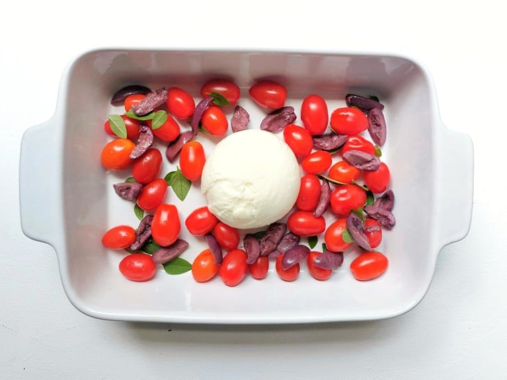 Um refratário contendo uma burrata, tomates-cereja, azeitonas picadas, salsinha e manjericão.