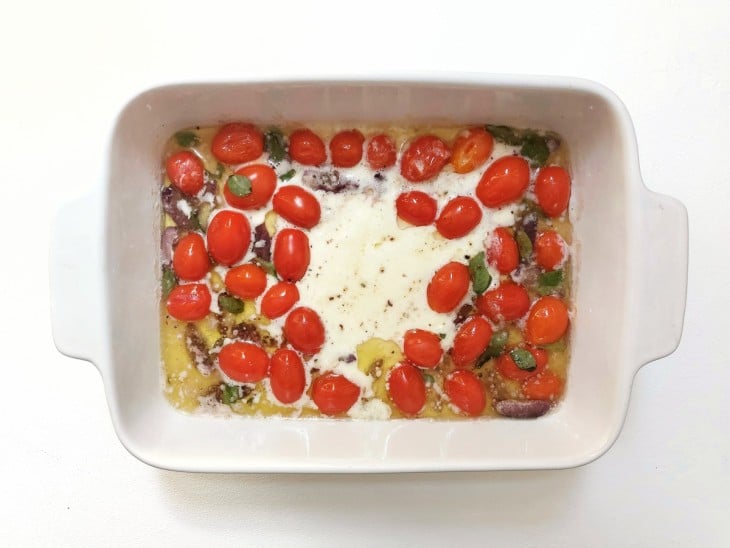Um refratário contendo burrata derretida, tomates-cereja, azeitonas picadas, salsinha e manjericão.