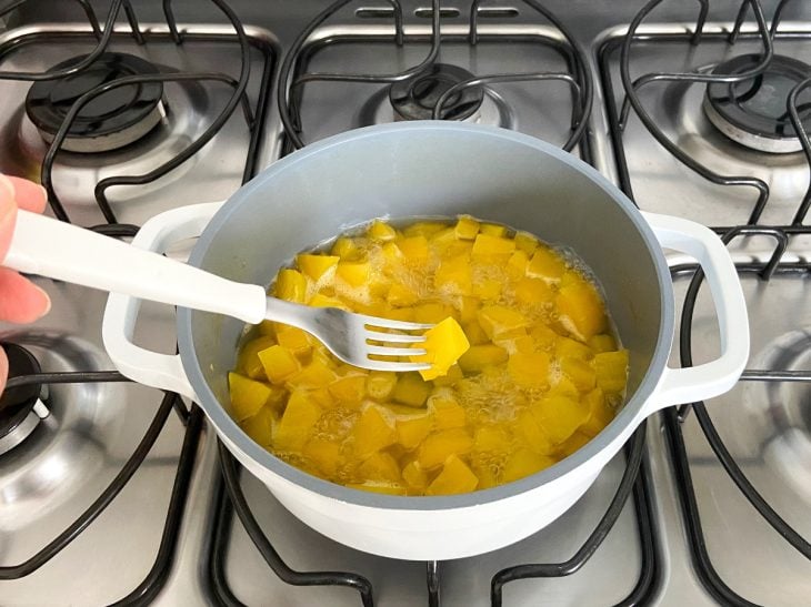 Uma mão com um garfo conferindo o ponto da mandioquinha na panela com água.