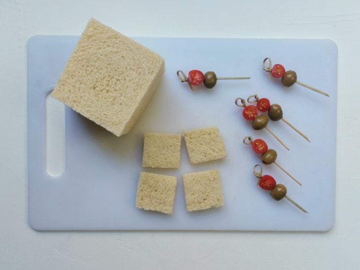 Uma tábua contendo fatas de pão de forma sem casca e palitinhos com tomate-cereja e azeitonas espetados.