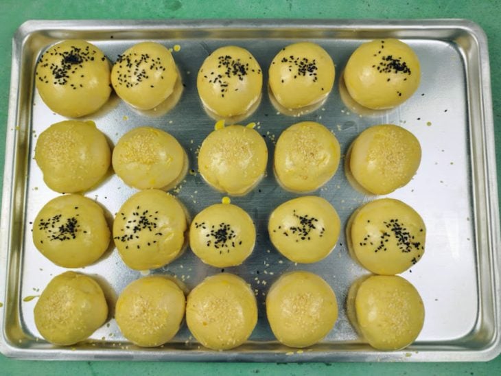 Uma assadeira com os pães pincelados com gema e leite, e gergelim salpicado por cima.