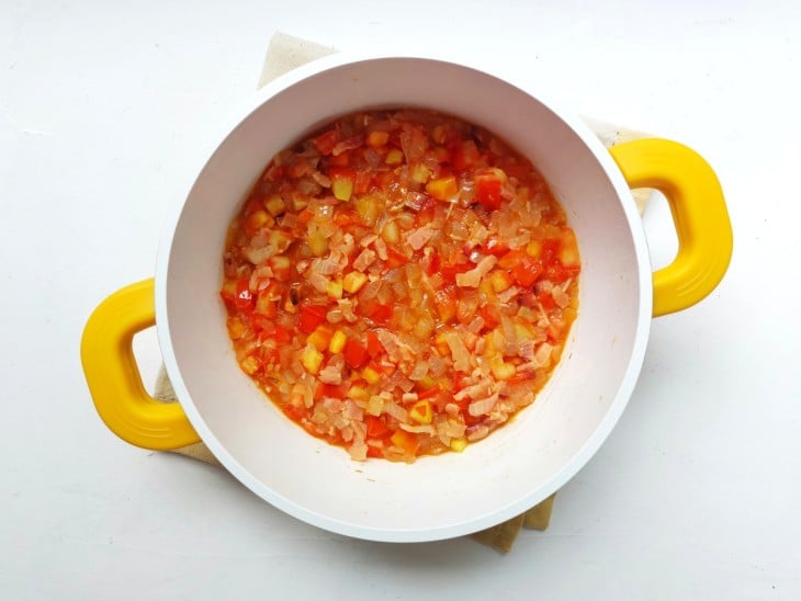 Tomates e cebolas adicionados a mistura de bacon, azeite e alho, na panela de cor branca e alças de cor amarela.