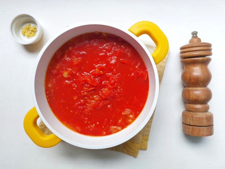 Uma panela na cor branco com alças na cor amarela, repleta de um molho com a coloração avermelhada. Ao lado, pimenta e gengibre em recipientes diferentes.