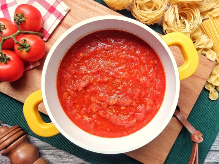 Molho à matriciana pronto dentro de uma panela de cor branca com alças de cor amarela. Ao lado, alguns tomates e massas cruas para decorar o ambiente.