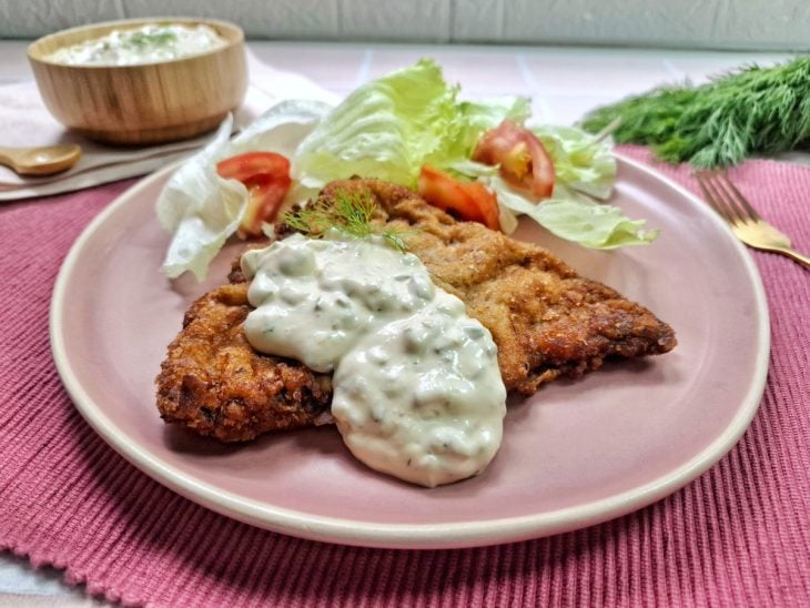 Molho tártaro servido com frango empanado e salada de alface e tomate em um prato rosa claro.