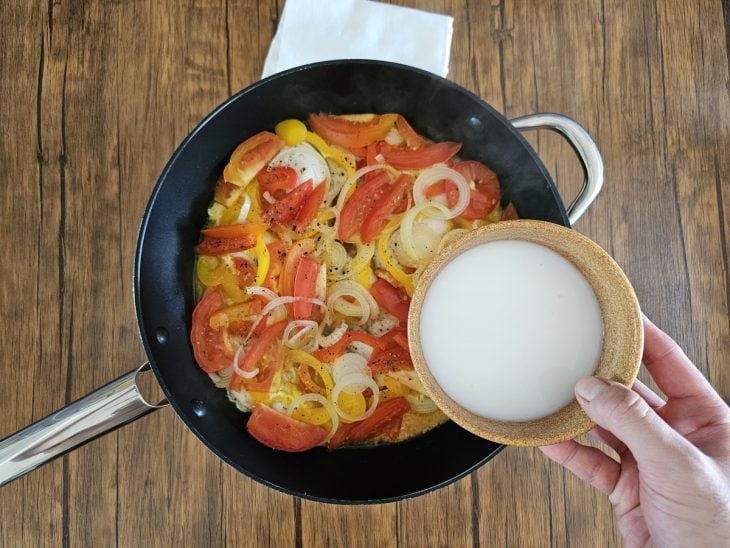 Uma panela cozinhando camadas de pimentões, cebola, postas de peixe, cebola e tomate com leite de coco.