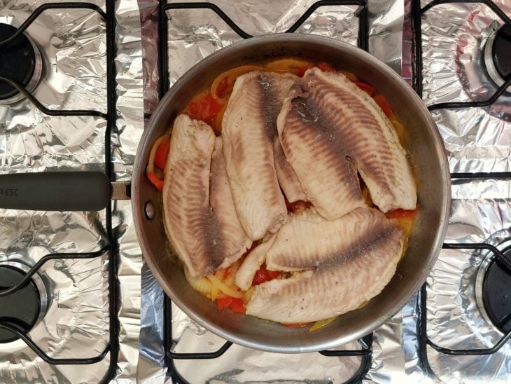 Uma frigideira refogando cebola, pimentão amarelo, pimentão vermelho, cebola e peixe.