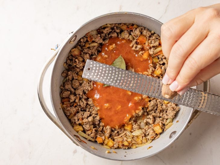 Panela com molho de tomate, folha de louro e noz-moscada sendo ralada.