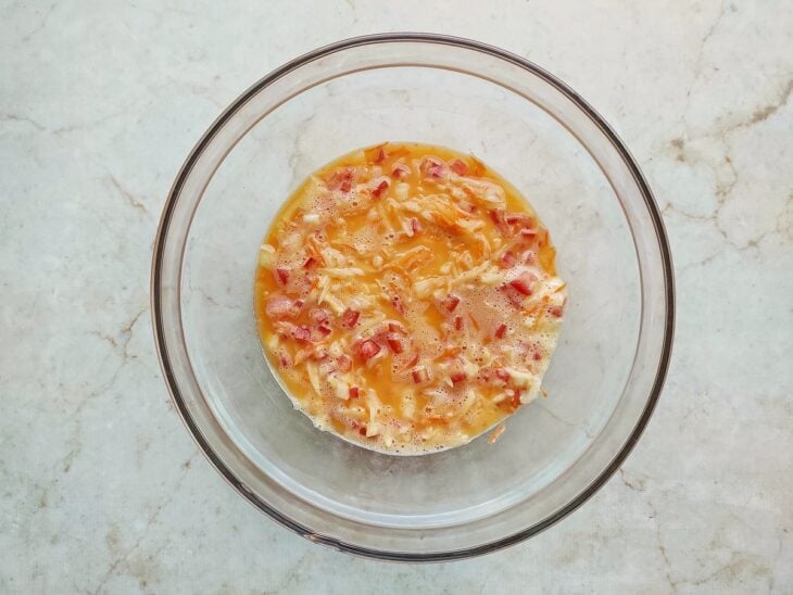 Cebola picada, tomate em cubos, batata e cenoura raladas sendo despejados nos ovos.