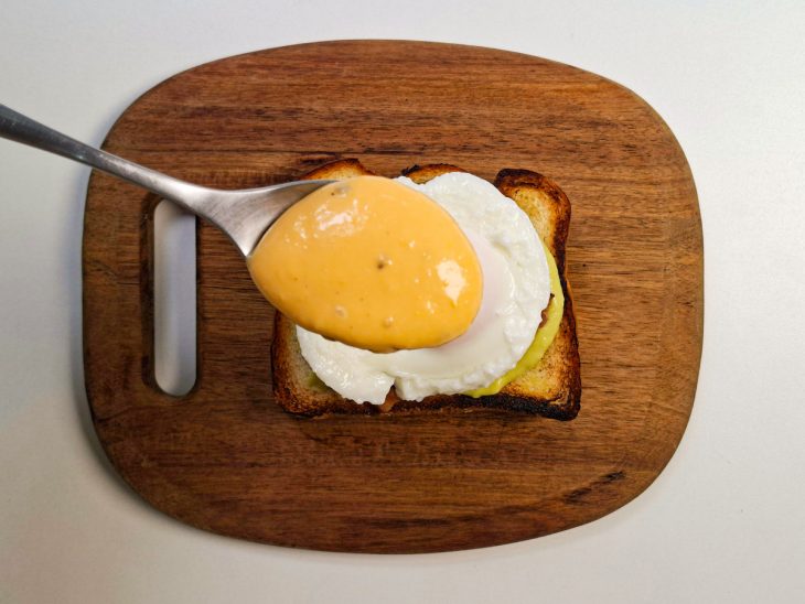 Ovo acomodo sobre o bacon e molho sendo despejado com auxílio de uma colher.