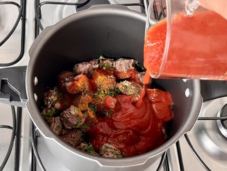 Uma panela de pressão contendo cubos de carne selados, temperos e molho de tomate.