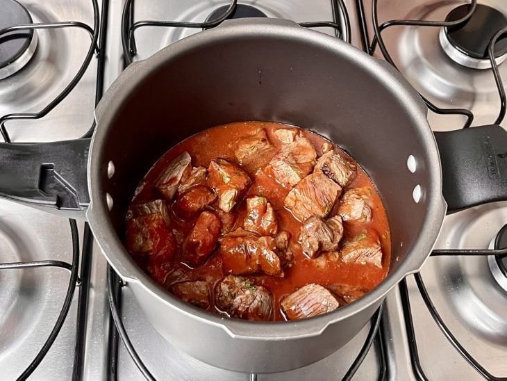 Uma panela de pressão contendo cubos de carne selados, temperos, molho de tomate e água.