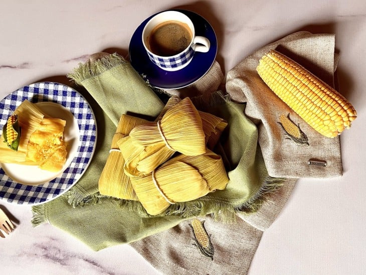 No centro, pamonhas nas trouxinhas de de palha amarradas com barbante. Ao lado, um prato com a pamonha aberta. Decoração com milho e servido com café.