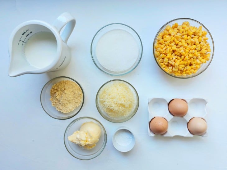 Ingredientes reunidos em diferentes recipientes: ovos, milho, leite, açúcar, manteiga, parmesão ralado, fubá e fermento.