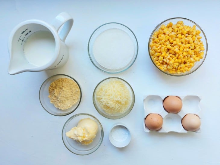 Ingredientes reunidos em diferentes recipientes: ovos, milho, leite, açúcar, manteiga, parmesão ralado, fubá e fermento.