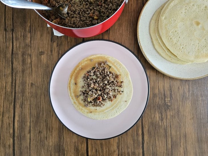 Uma massa de panqueca aberta sendo recheada com carne moída.