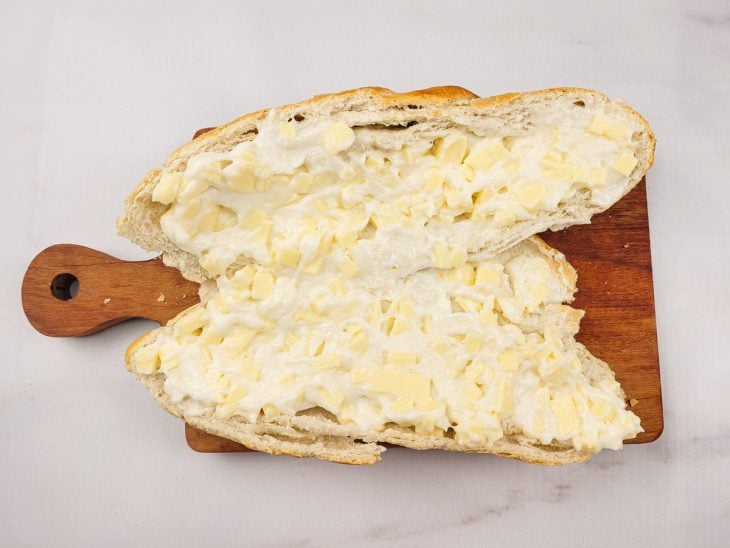 Uma tábua contendo um pão italiano aberto com a mistura de mussarela e requeijão.