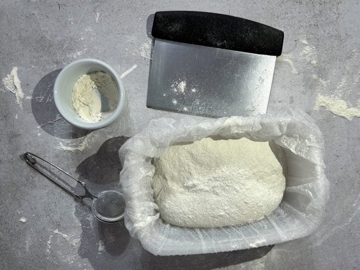 Massa acomodada em um recipiente forrado com plástico, acompanhada de farinha e cortador.