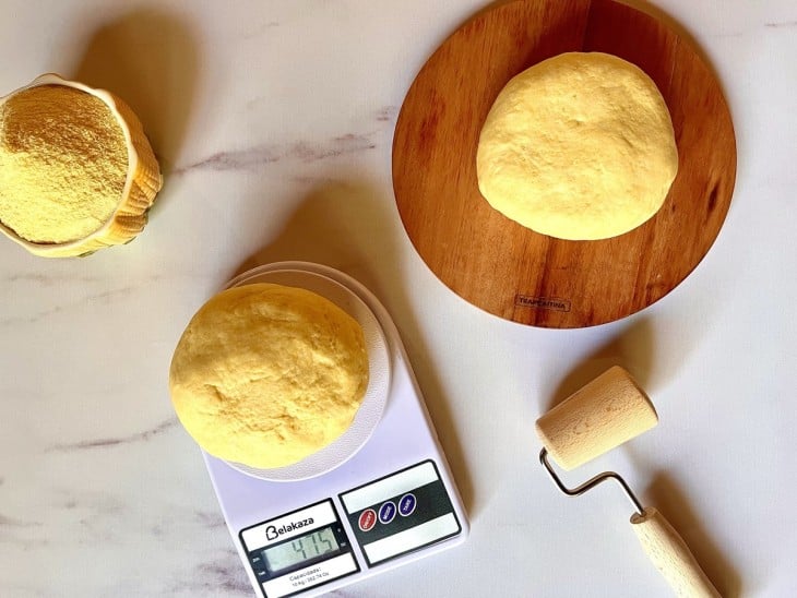 Massa do pão de cor amarelada dividida em duas partes, uma delas em cima de uma balança. Um recipiente ao lado cheio de fubá.