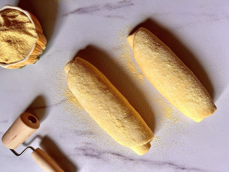 As duas massa de pão enroladas em formato de rocambole e passadas no fubá, dispostas em cima de uma bancada.