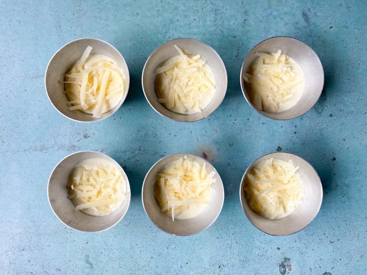 Recipientes contendo massa de pão de queijo cobertos com queijo ralado.