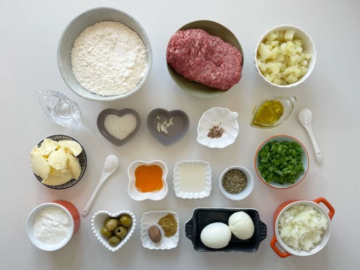 Ingredientes organizados sobre a bancada com legumes, carne, farinha e demais itens separados para o preparo.
