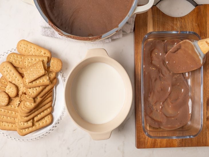 Bolachas, creme de chocolate em uma panela e espalhado em uma travessa, e tigela com leite para umedecer as bolachas, reunidos em uma bancada.