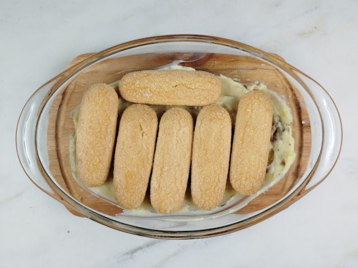 Biscoitos champagne umedecidos com leite posicionados sobre a camada de creme de nozes no refratário.