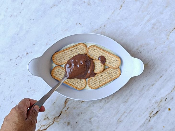 Camadas sendo montadas na travessa com creme branco, bolacha e creme de chocolate.
