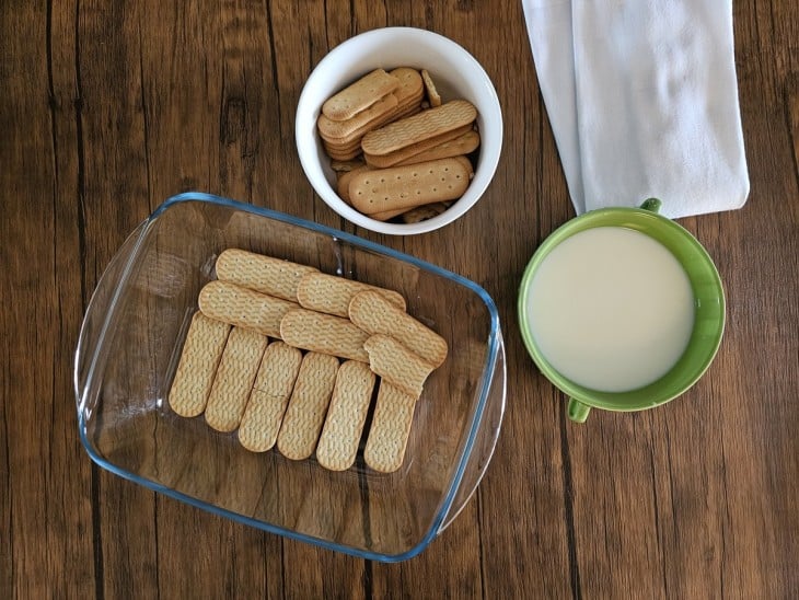 Camada de bolachas em uma travessa, potinho com bolachas e leite em uma vasilha.