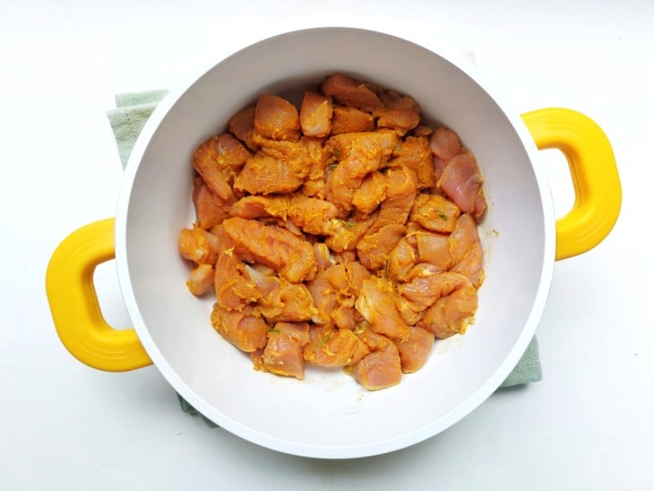 Carne em uma coloração alaranjada dos temperos, dentro de uma panela branco com alças em amarelo.