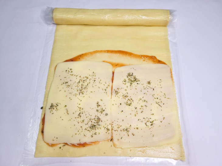 Uma massa de pastel aberta contendo molho de tomate espalhado, com uma camada de mussarelas com orégano.