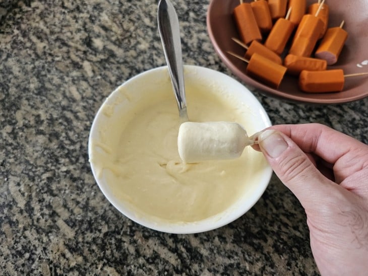 Uma salsicha espetada sendo passada na massa líquida.