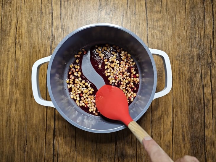 Mexendo com uma espátula parte da pipoca com corante vermelho dentro de uma panela cinza com as alças brancas