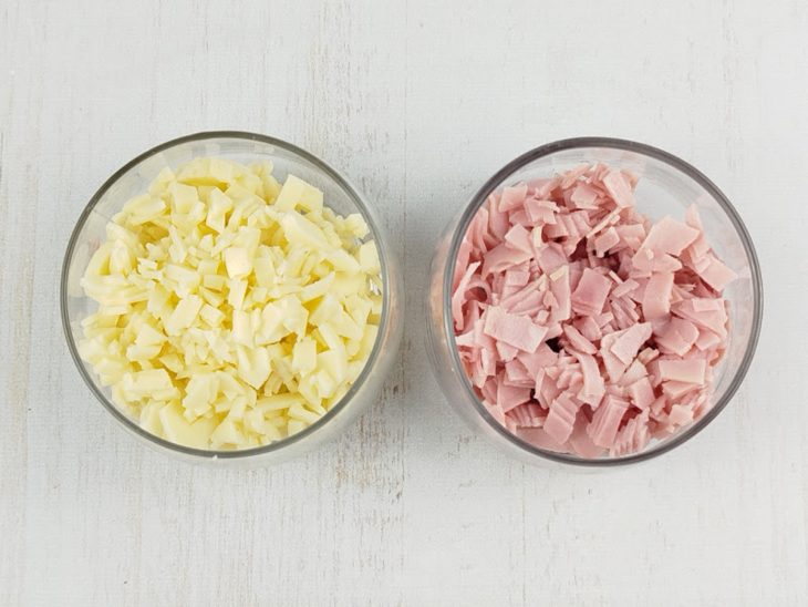 Queijo e presunto picados e reservados em recipientes redondos de vidro.