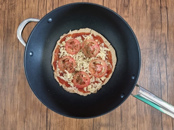 Recheios de tomate, queijo e molho sobre pizza, dentro de frigideira preta.