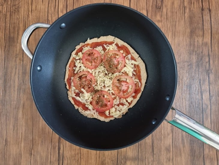 Recheios de tomate, queijo e molho sobre pizza, dentro de frigideira preta.
